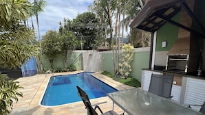 A swimming pool with a glass table and chairs, surrounded by greenery and a barbecue area.