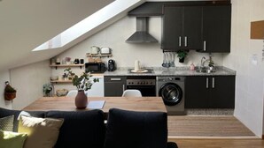 A compact kitchen with dark cabinets, a microwave, a stove, and a washing machine. There's a dining area with a table and chairs, and a small plant on the counter. - Apartamento El Picu (Collanzo)