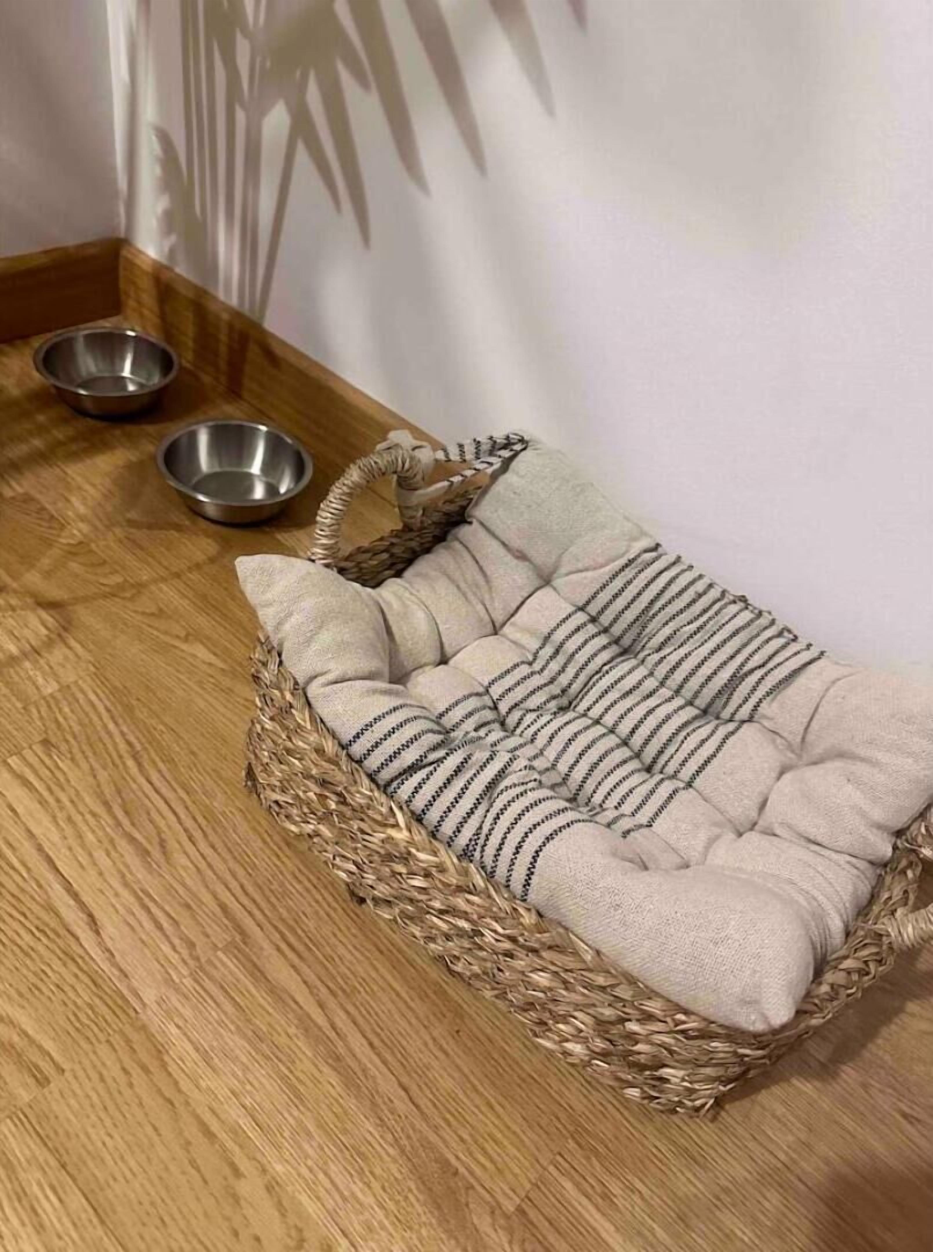 A pet bed with a woven basket base and striped cushion, placed on a wooden floor next to a wall with a shadow of a plant.