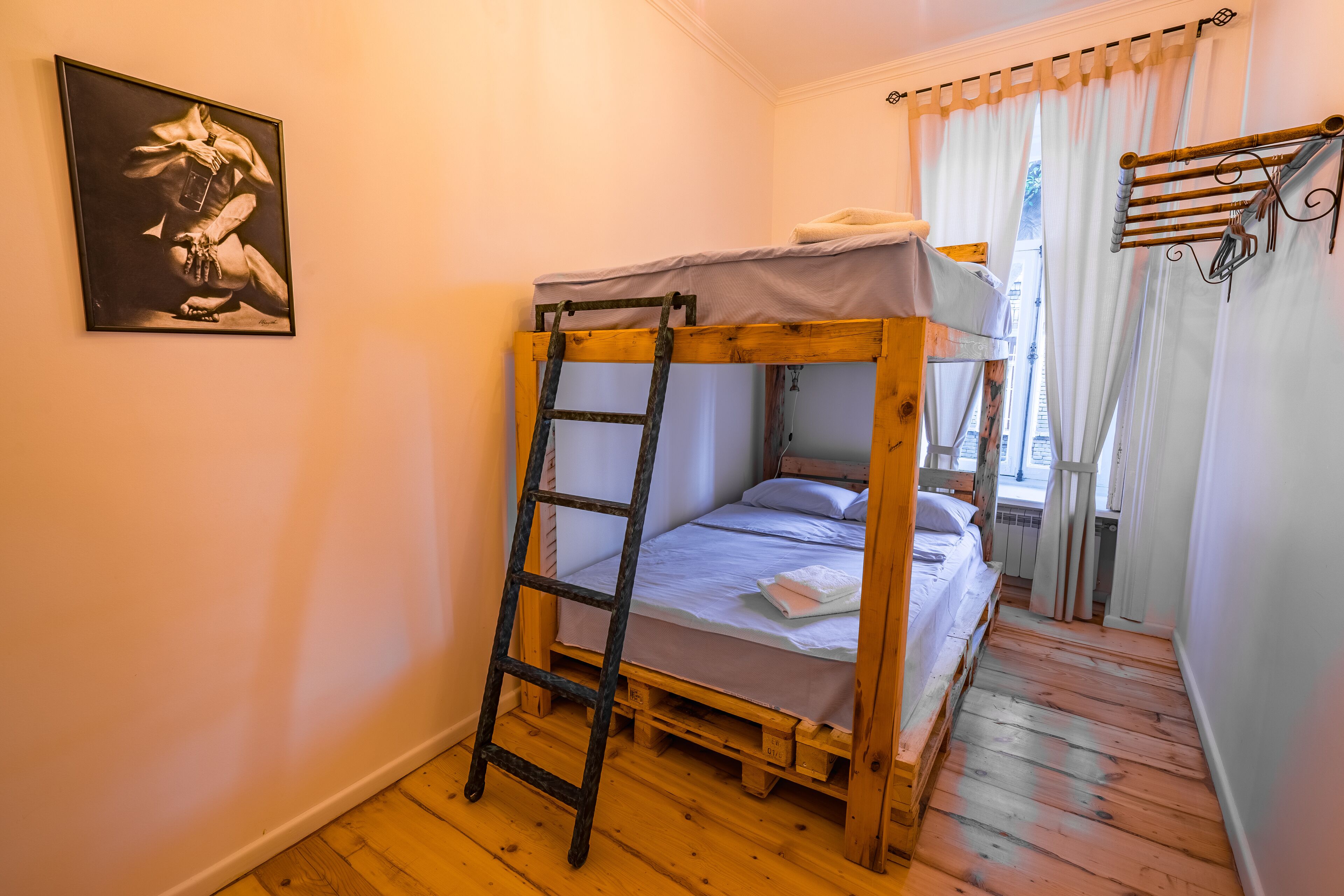 A bunk bed with a ladder, a wooden floor, a window with curtains, and a wall-mounted towel rack.