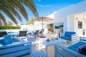 Zona d'estar exterior moderna amb mobles blancs, una piscina i una porta de vidre que dona a una habitació.