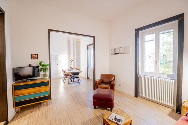 A living room with a wooden floor, a TV on a wooden cabinet, a leather armchair, and a dining area with a table and chairs. - Charming Design Apartment in Vibrant Pigneto (Roma)