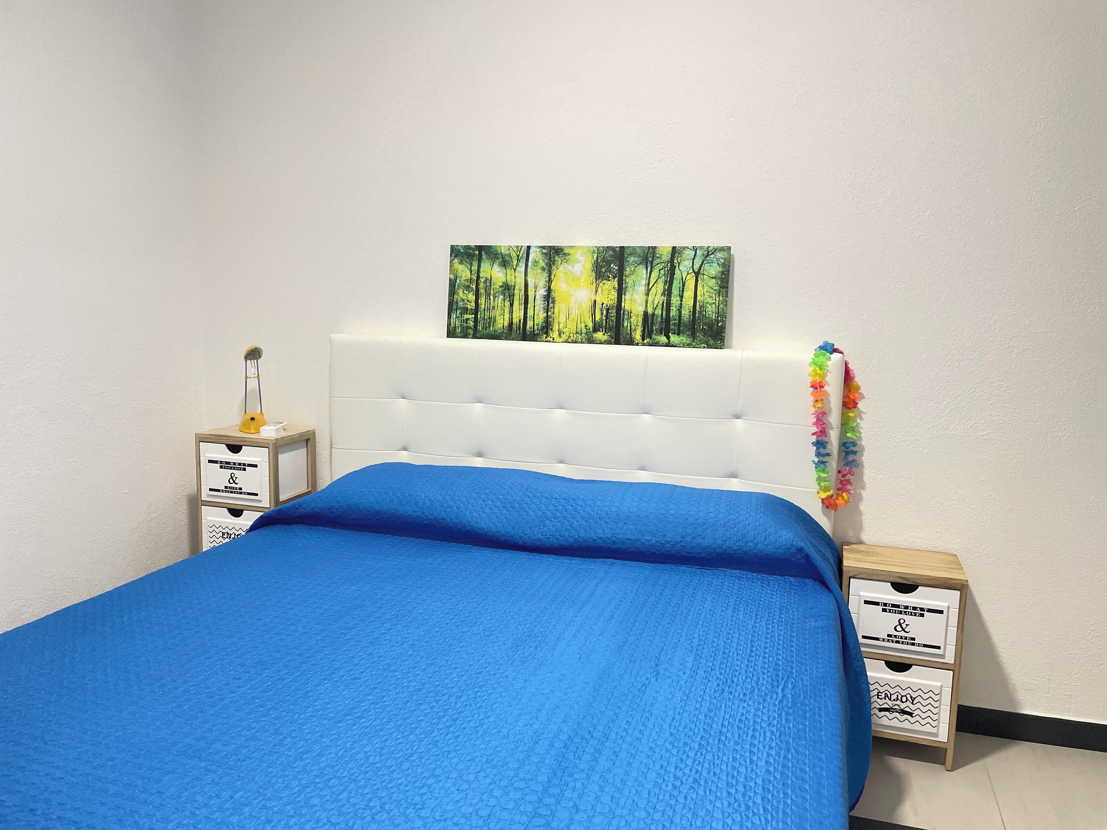 A bed with a blue bedspread, a white headboard, and a forest-themed painting above it. There are two nightstands with drawers and a small lamp on one.