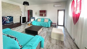 Una sala de estar moderna con un sofá, una mesa de centro y un televisor. Hay una decoración con forma de corazón rojo en la pared y una ventana con cortinas.