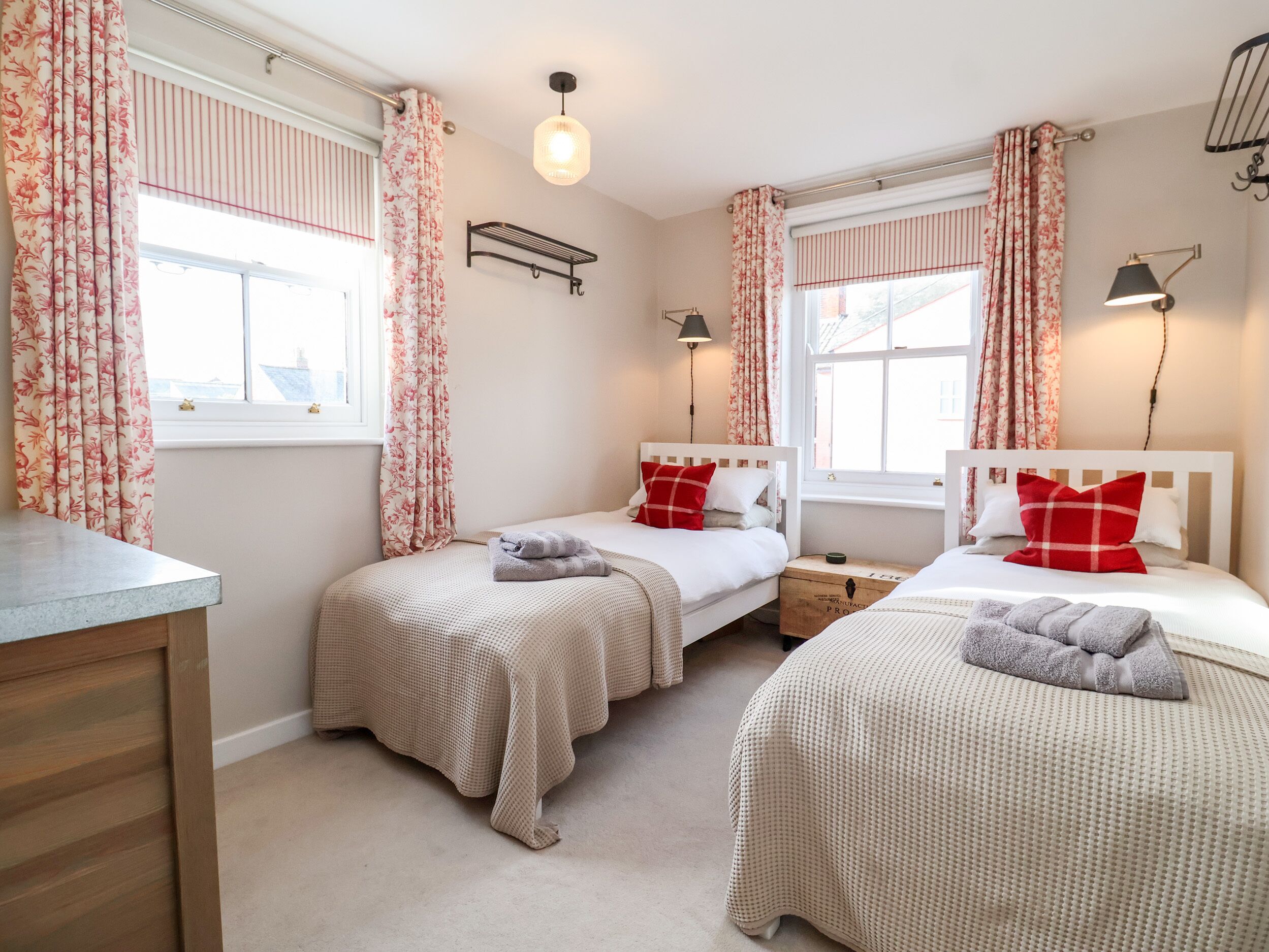 A room with three beds, each with a white headboard and red pillows, a small wooden nightstand, a wall-mounted shelf, and a window with striped curtains.