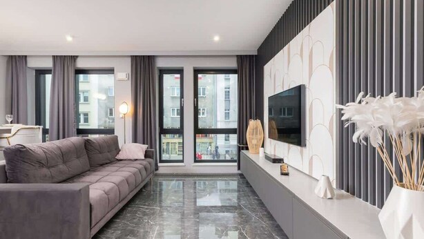 Una sala moderna con un sofá gris, un televisor de pantalla plana montado en la pared y un amplio ventanal con cortinas.