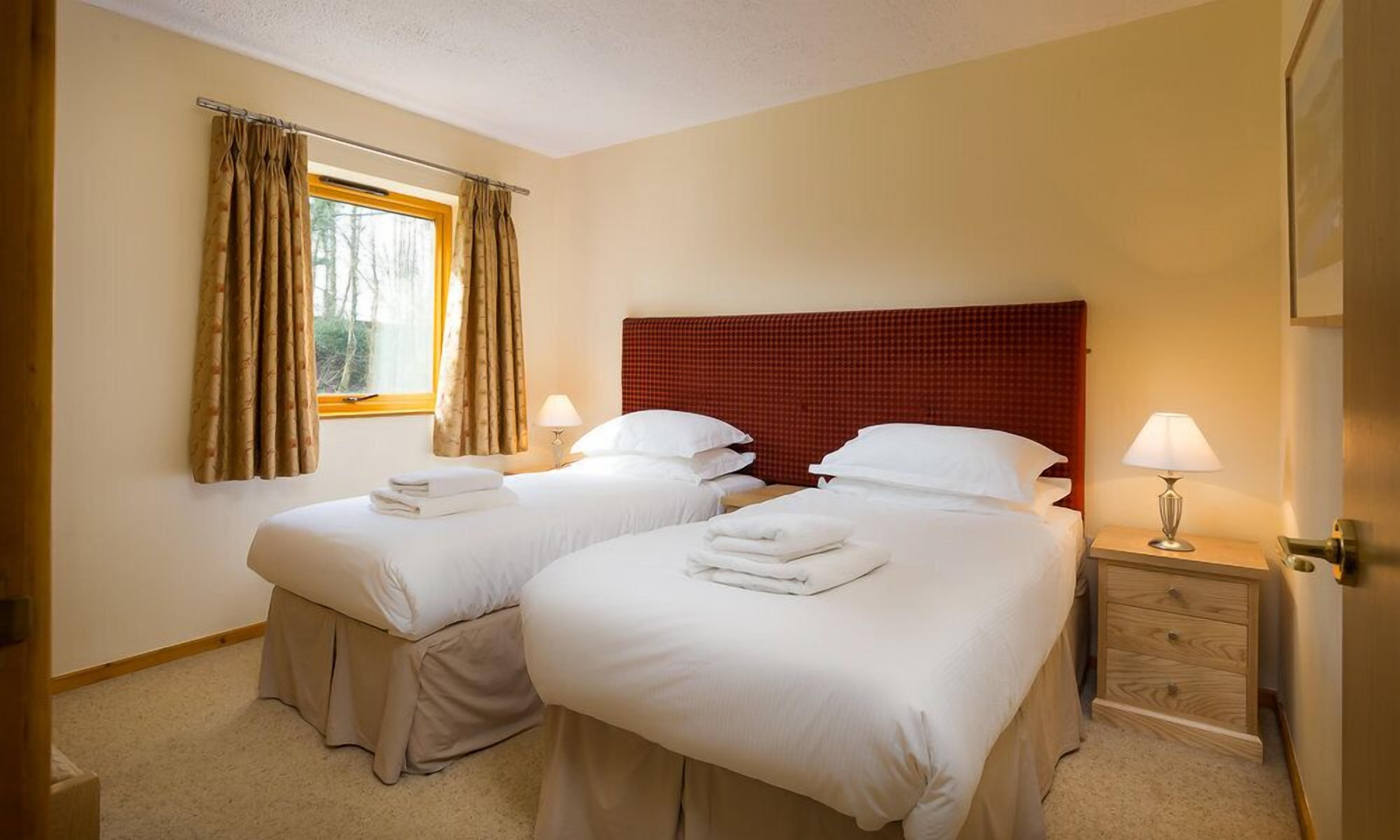 Ein Hotelzimmer mit zwei Betten, einem Fenster mit Vorhängen, einem Nachttisch mit Lampe und einem Holzkoffer.