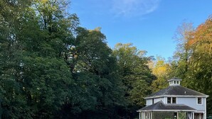 Une maison à deux étages, dotée d’un toit à pignon et d’un petit porche, entourée d’arbres denses et sous un ciel dégagé.