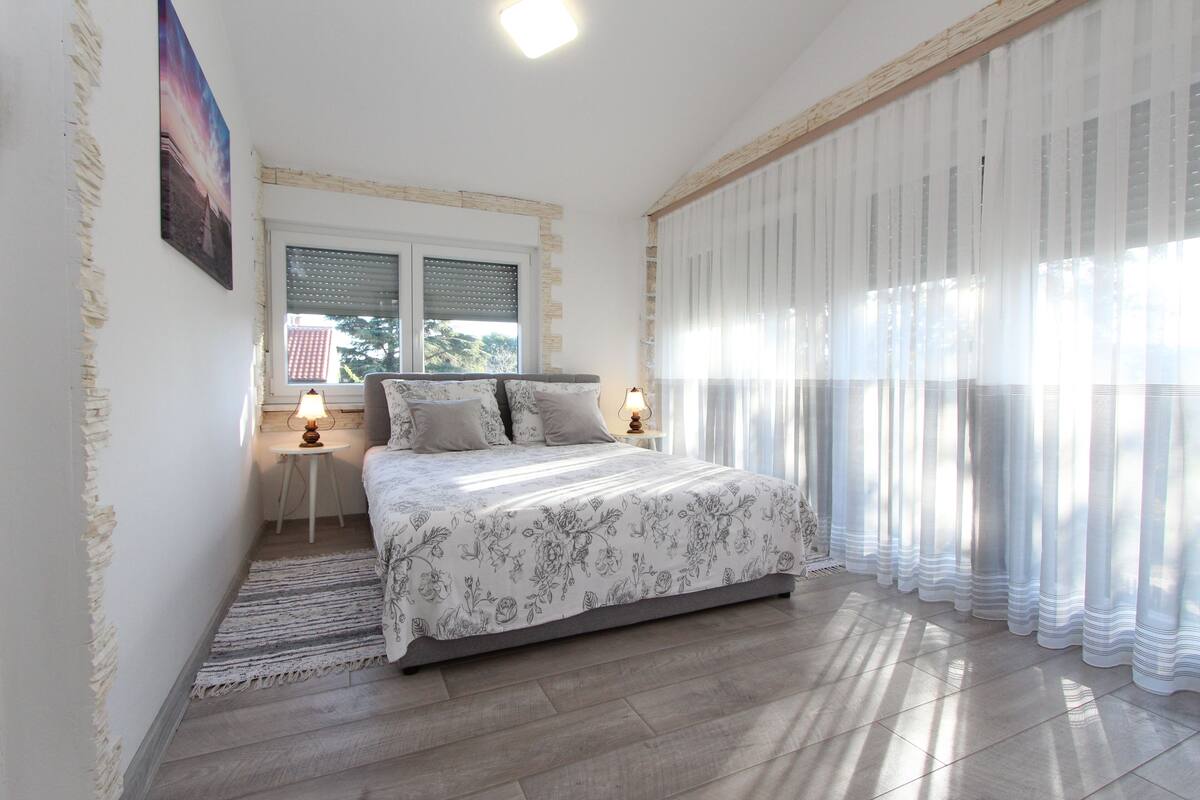 A bedroom with a bed, a window with blinds, a nightstand with a lamp, and a view of the outside.