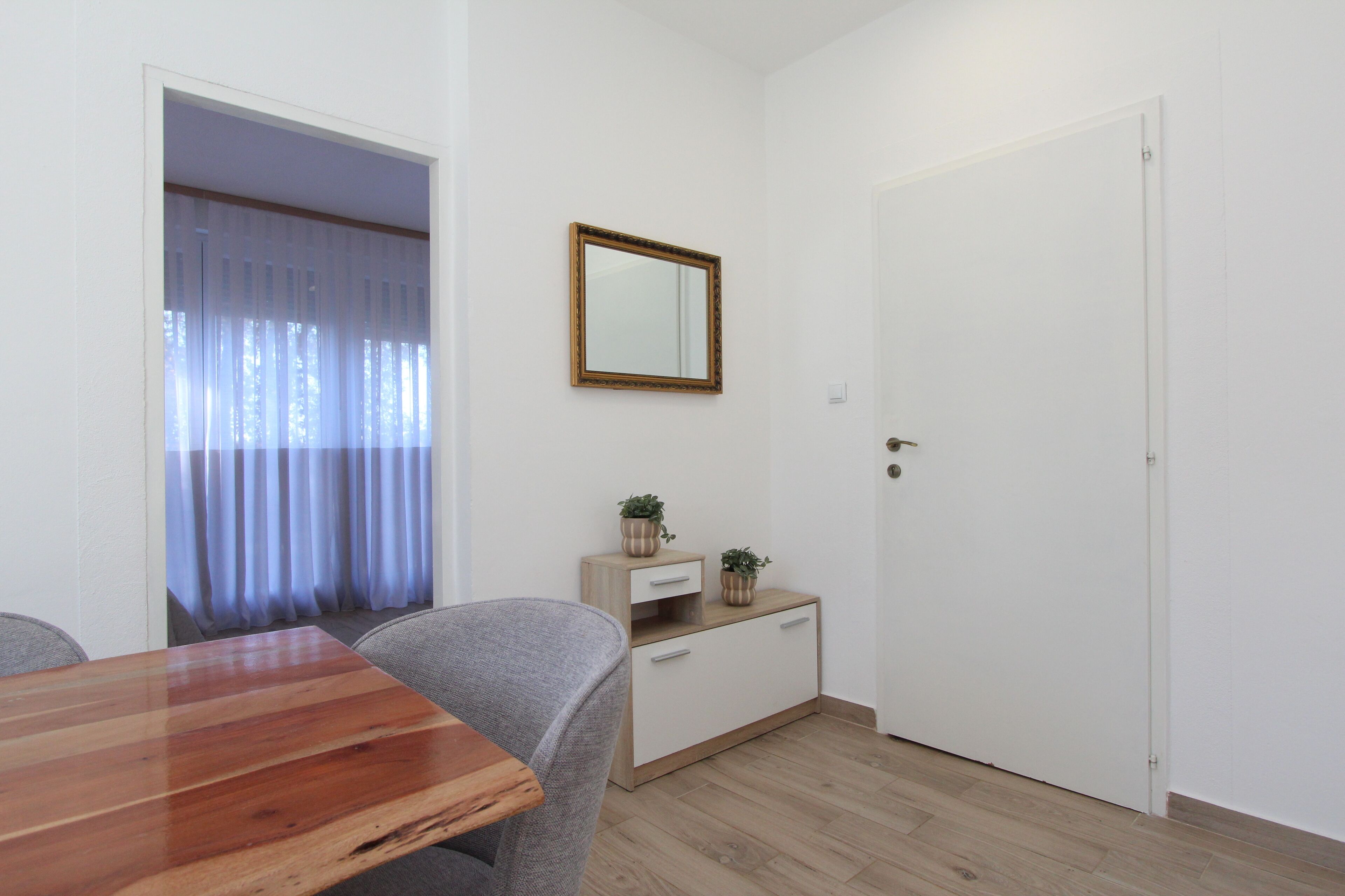 A room with a wooden dining table, grey upholstered chairs, a small white cabinet with two potted plants, a mirror on the wall, and a white door.