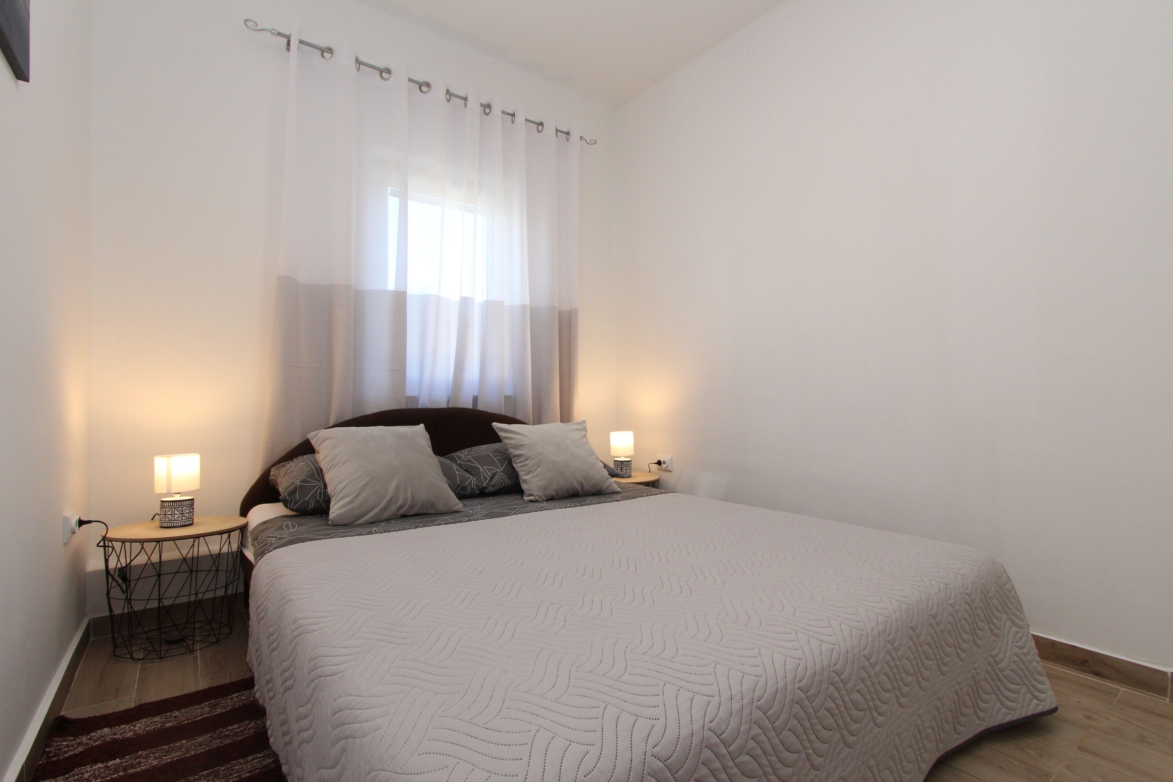 Un lit bien fait, recouvert d’une couvre-lit à motifs, avec deux lampes de chevet et une fenêtre ornée de rideaux légers.