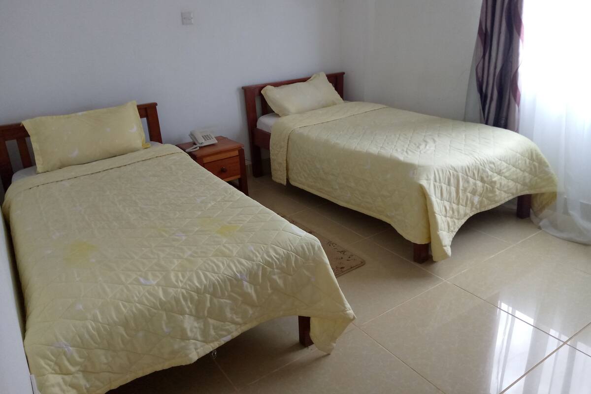 Two single beds with matching yellow bedding, a wooden nightstand with a telephone, and a tiled floor.