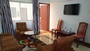 Ein Hotelzimmer mit braunen Sofas, einem Glastisch, einem Holzstuhl, einem kleinen Tisch mit Flasche und Tasse sowie einem Fernseher an der Wand.