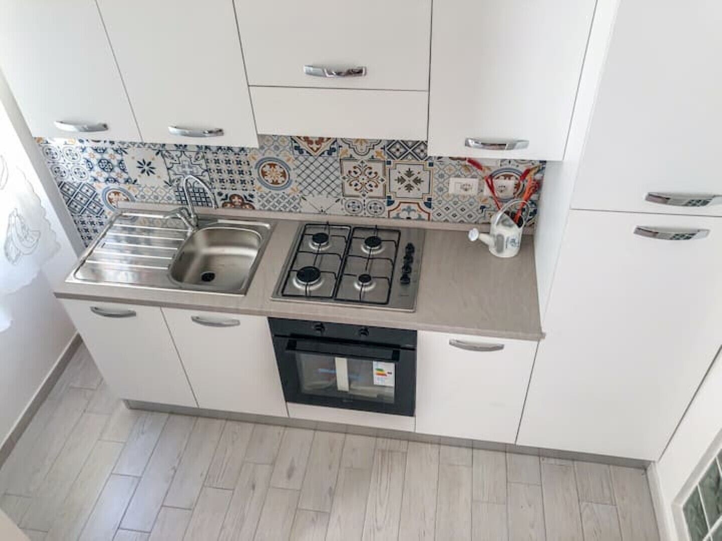 Uma cozinha compacta com armários brancos, pia de aço inoxidável, fogão a gás e lava-louças. O revestimento da parede apresenta um padrão de azulejos coloridos.