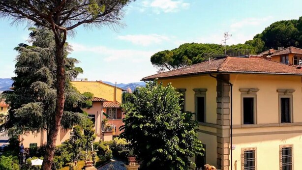 A traditional building with a yellow facade, a large tree in front, and a mountainous backdrop.