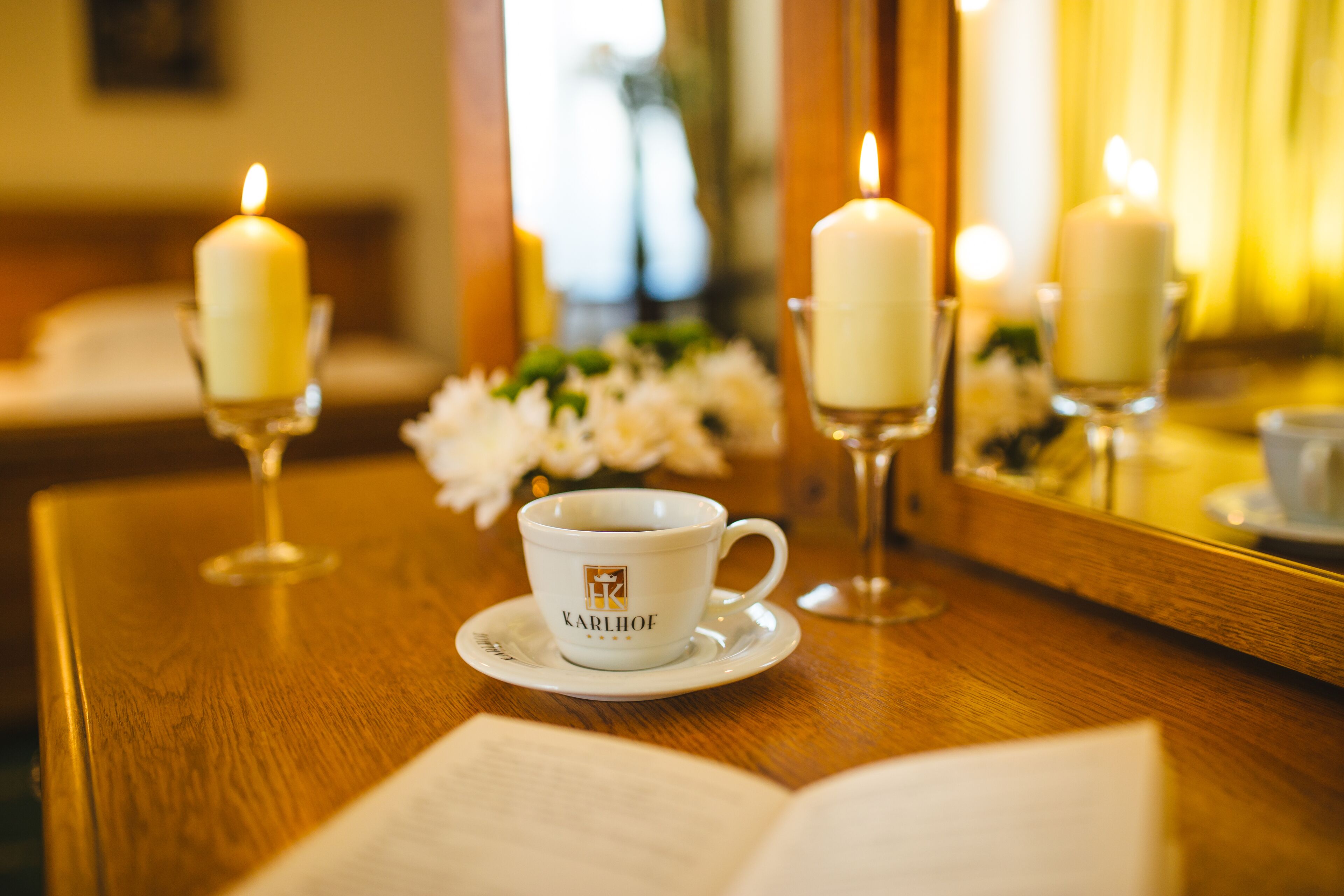 Une table sur laquelle se trouvent une tasse blanche portant l’inscription « KARLHOFF » et un livre, le tout entouré de bougies et d’un miroir.