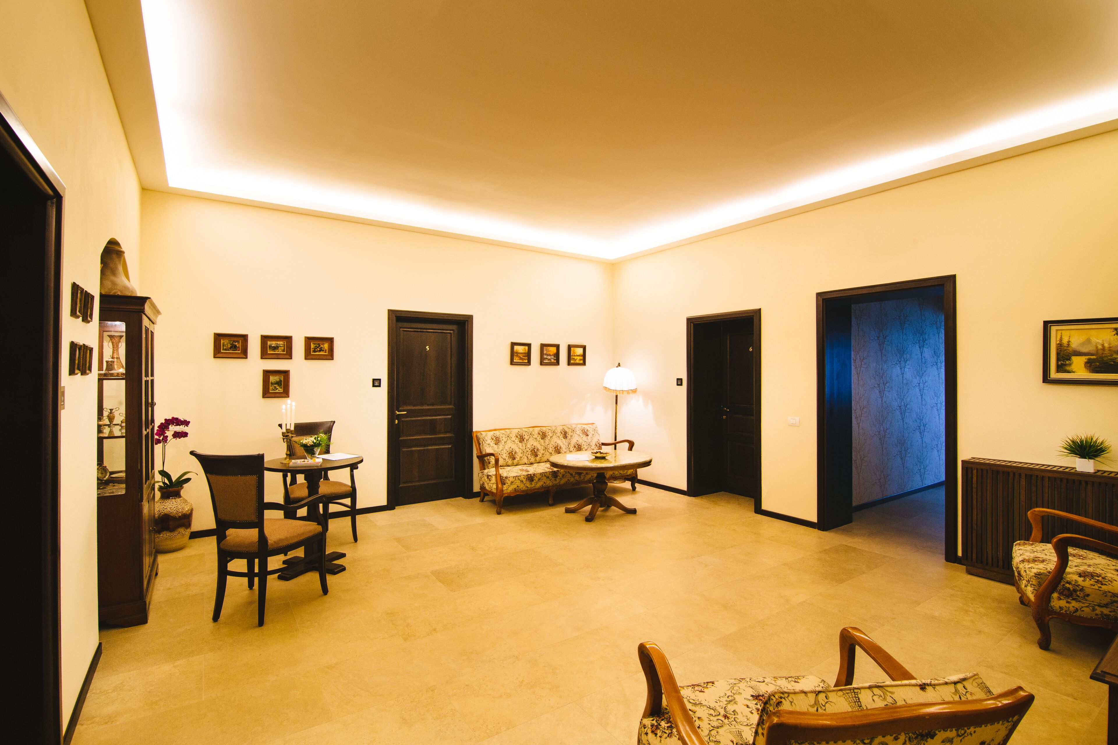 A spacious living room with a sofa, chairs, a coffee table, and a lamp. There are framed pictures on the walls and a door leading to another room.