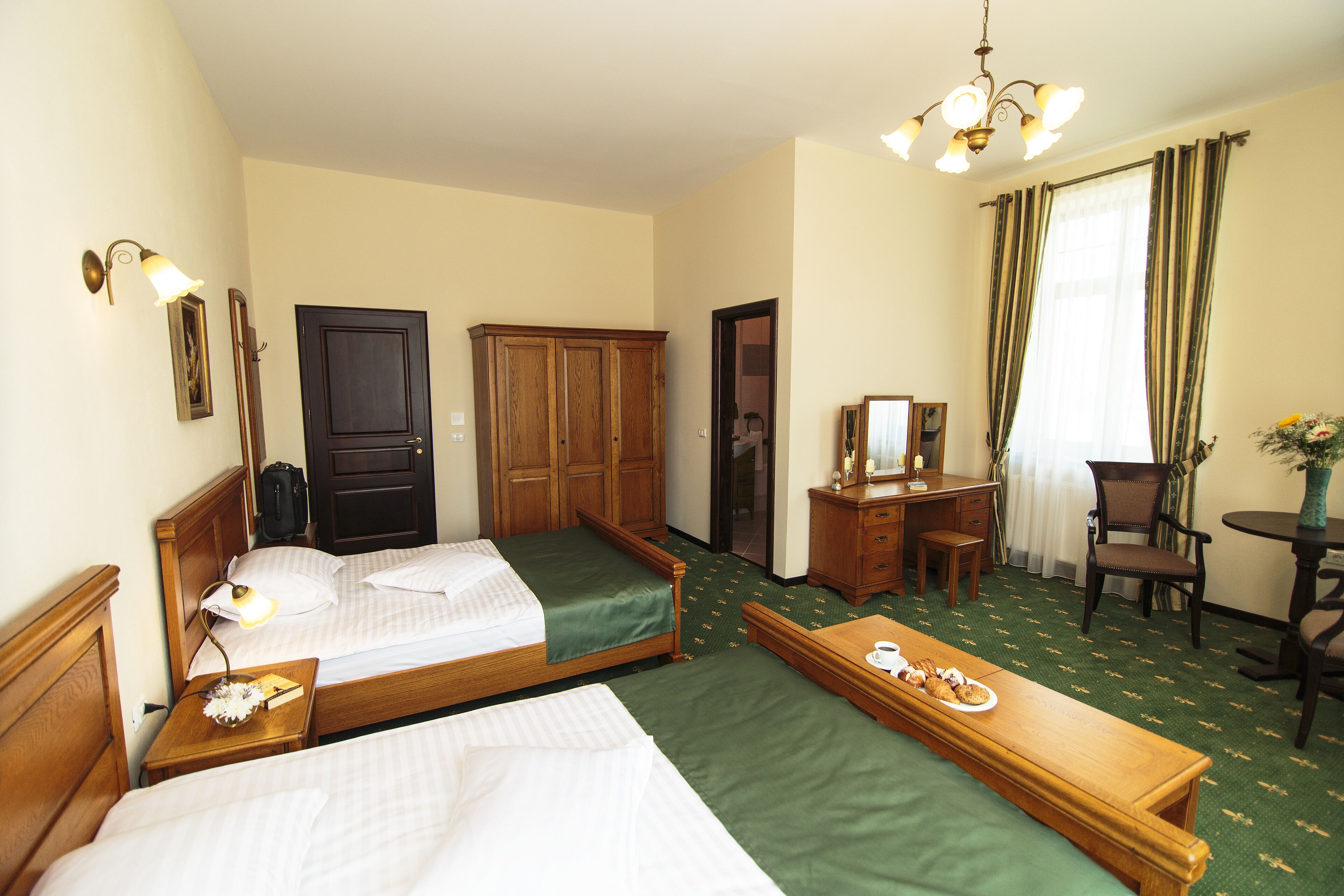 A hotel room with two single beds, a wooden desk, a chair, a small table with a plate of food, a mirror, and a lamp.