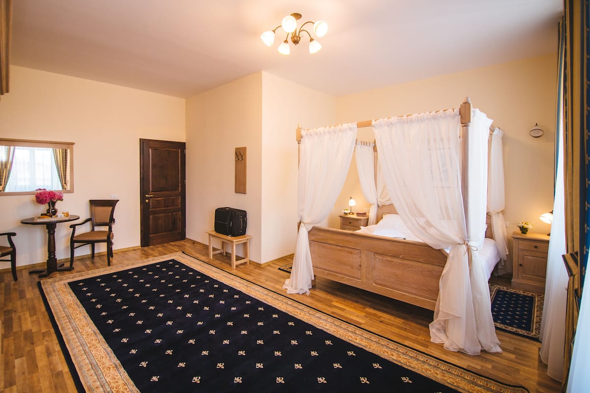 Une chambre Ă coucher avec un lit Ă baldaquin, une commode en bois, une table de chevet, une petite table avec une chaise et un grand tapis Ă motifs.