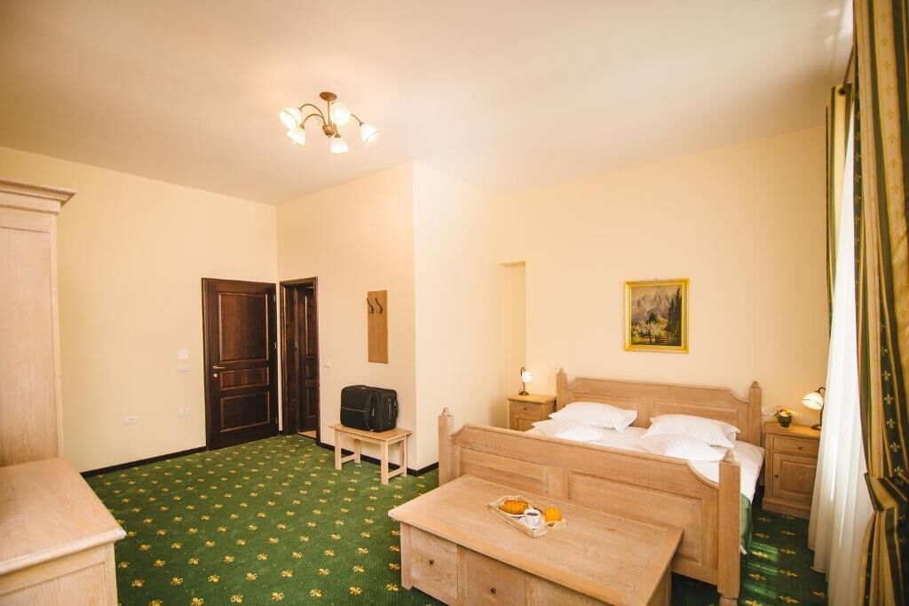 A hotel room with a green patterned carpet, a wooden bed, a small bedside table with a lamp, and a painting on the wall.