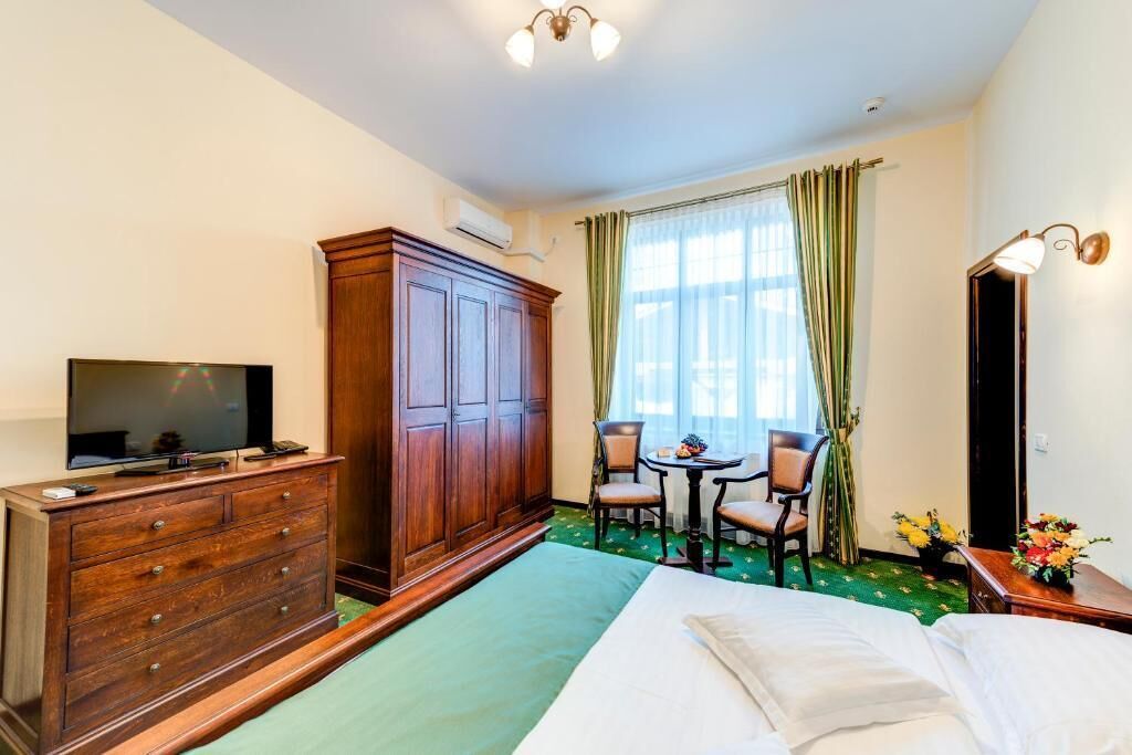 Une chambre d’hôtel comprenant un lit, une commode en bois, un téléviseur, un bureau avec deux chaises, une fenêtre avec des rideaux et un luminaire fixé au mur.
