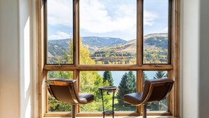 Ein Zimmer mit großem Fenster und Blick auf eine Berglandschaft. Zwei Stühle und ein kleiner Tisch stehen im Raum.