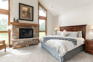 Une chambre avec une cheminée en pierre, de grandes fenêtres, un lit recouvert d’une couvre-lit à motifs et une table de chevet avec une lampe.