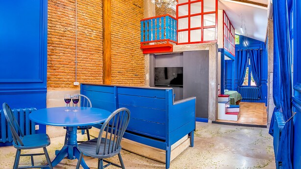 Ein Zimmer mit blauem Esstisch und Stühlen, einem blauen Sofa, einer Ziegelwand und einer Treppe mit blauen Geländern.