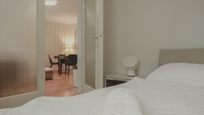 Ett hotellrum med en säng, ett nattduksbord med en lampa och en dörr som leder till en matplats.