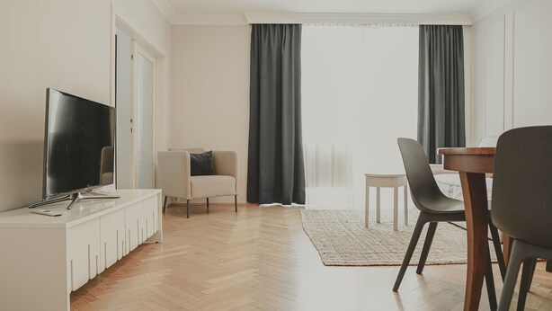 Ett modernt vardagsrum med en platt-TV, en vit TV-möbel, en soffa, ett matbord med stolar och stora fönster med mörka gardiner.