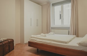 Une chambre à coucher avec un lit en bois, du linge de lit blanc, une fenêtre avec des rideaux et un radiateur.