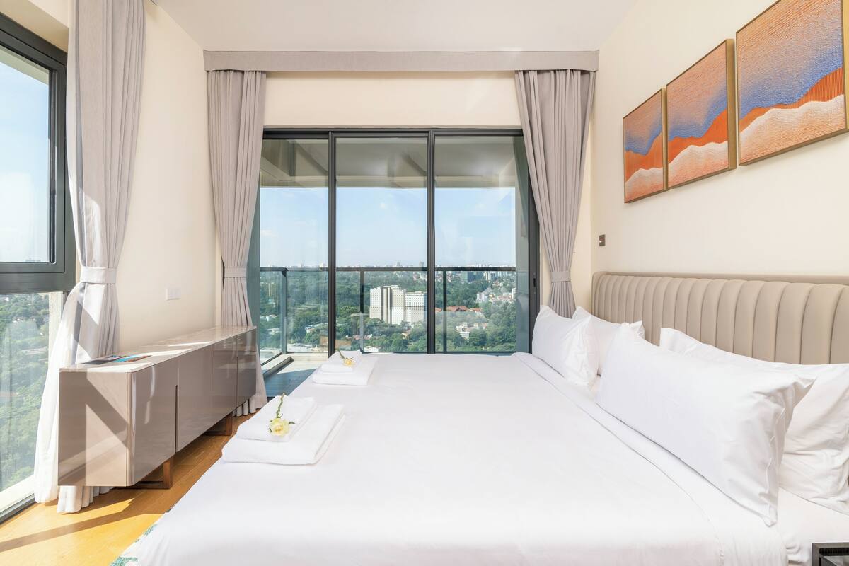 A bedroom with a large bed, a view of a cityscape, a window with curtains, and a painting on the wall.