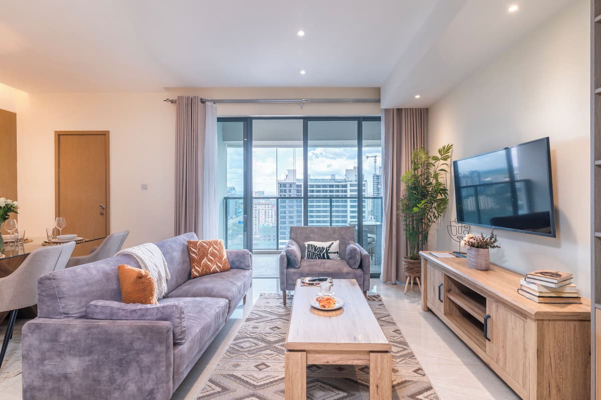A modern living room with a sofa, armchair, coffee table, TV, and a balcony with a city view.