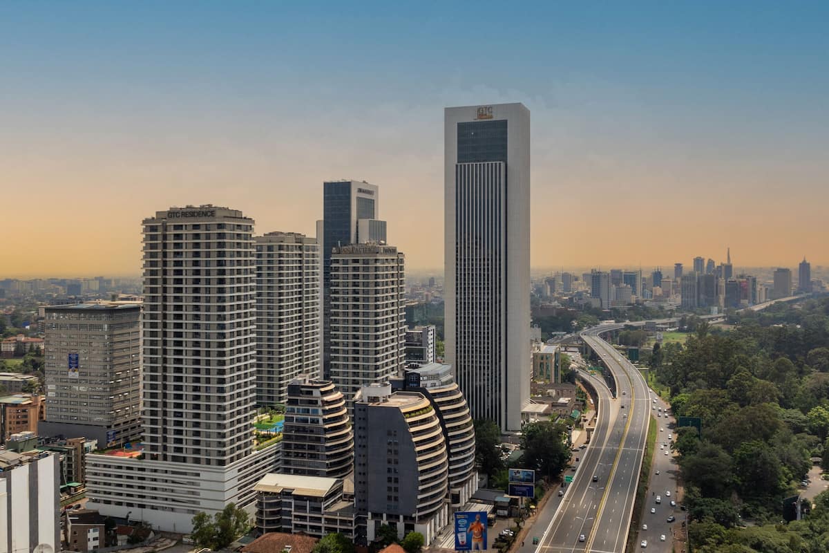 A high-rise building with a modern design, surrounded by other high-rise buildings, a curved highway, and greenery.
