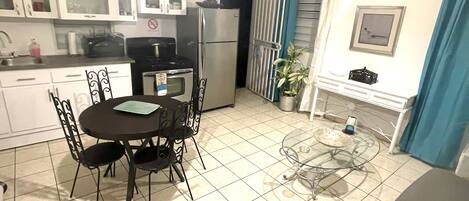 A compact kitchen with white cabinets, a dining table with chairs, a refrigerator, and a small potted plant.