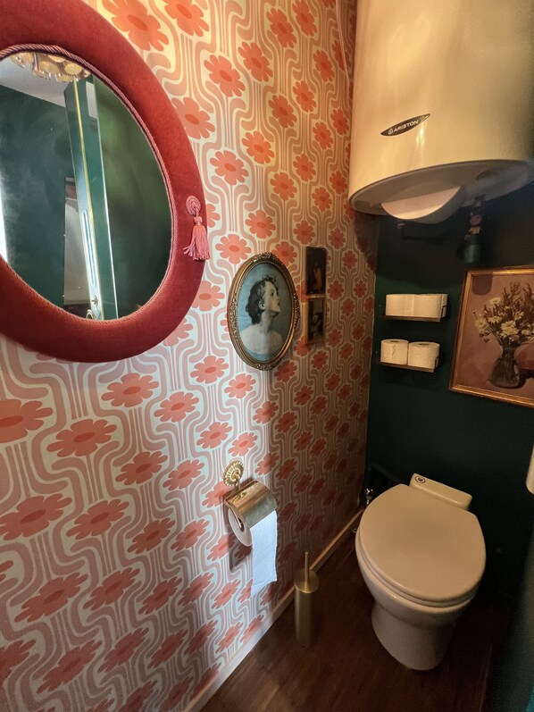 A bathroom with a patterned wallpaper, a round mirror, a framed picture, a toilet, and a roll of toilet paper on the wall.