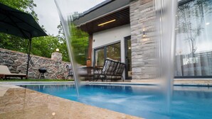 A modern outdoor pool area with stone walls, a stone grill, and a patio with chairs and an umbrella. - Vital Home (Sapanca)