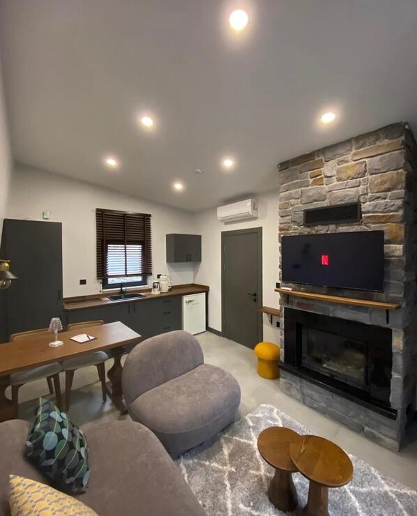 A living room with a stone fireplace, a television mounted above it, a dining area with a table and chairs, and a kitchen in the background.