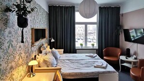 Ett hotellrum med en säng, ett skrivbord, en stol, en tv och ett fönster med gardiner.