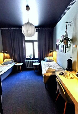 A compact hotel room with two single beds, a small table, a chair, a desk, a hanging light fixture, and a window with curtains. - Pks - Ät drick & Bo (Hudiksvall)