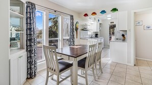 A dining area with a wooden table and white chairs, a kitchen in the background, and a view through the window.