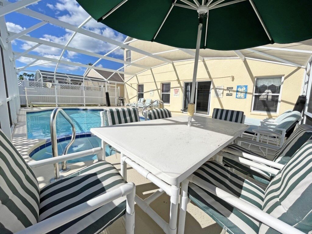 Un espace aménagé au bord de la piscine, avec une table blanche et des chaises à rayures, un parasol et une piscine.