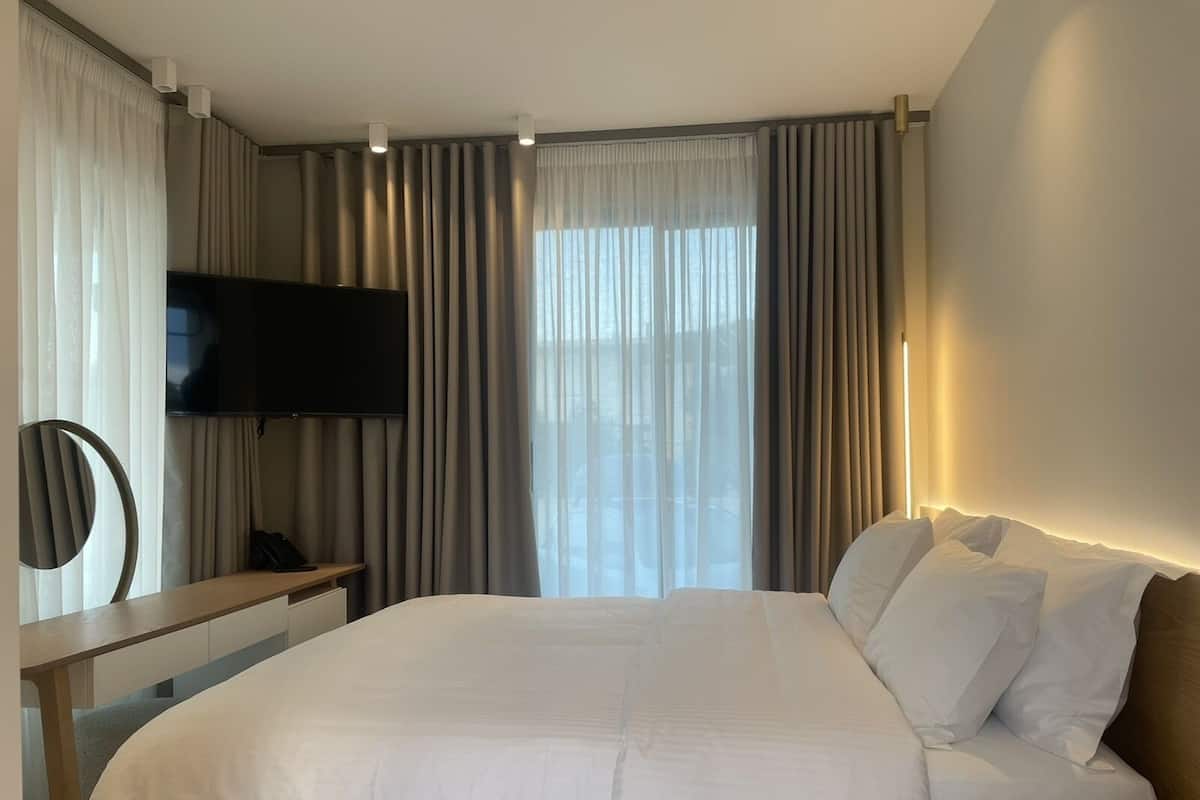 A modern hotel room with a large bed, a desk with a chair, a flat-screen TV, and a large window with curtains.