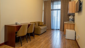 Een hotelkamer met een houten bureau, twee stoelen, een bank, een aan de muur bevestigde televisie en een kleine koelkast.