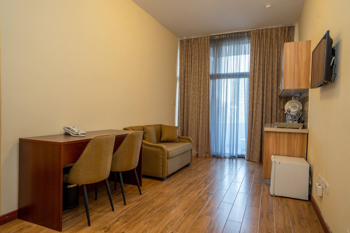 A hotel room with a wooden desk, two chairs, a sofa, a television mounted on the wall, and a small refrigerator.