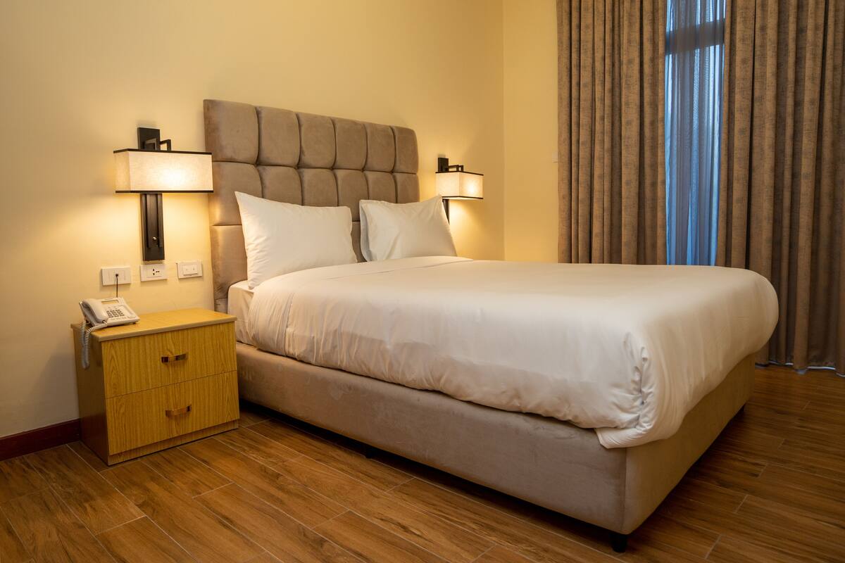 A hotel room with a large bed, two bedside lamps, a nightstand with a phone, and a window with curtains.