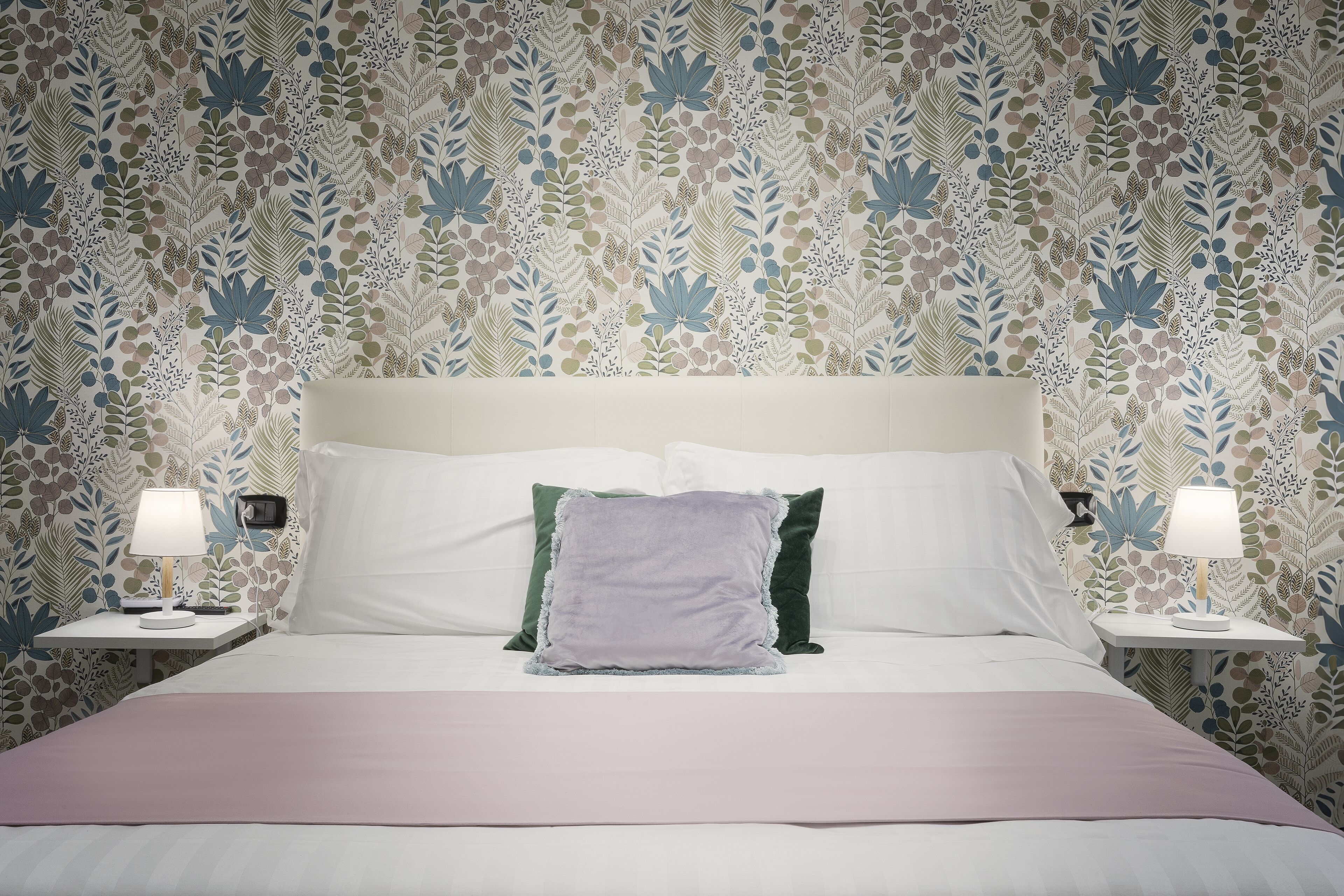 A neatly made bed with a pink bedspread, two bedside tables with lamps, and a floral patterned wallpaper.
