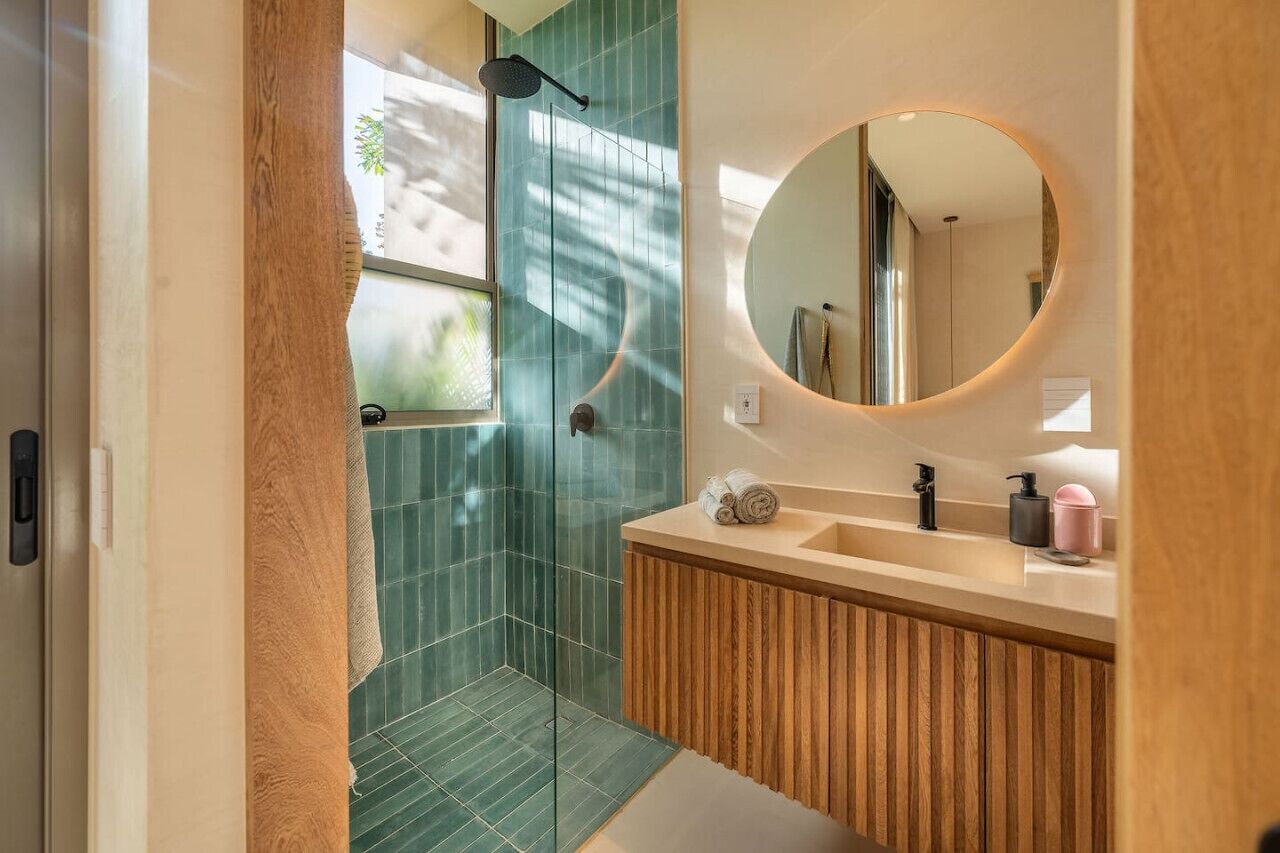 Ein Badezimmer mit einem Holzkosmetikschrank, einem runden Spiegel und einer Dusche mit grünen Fliesen.