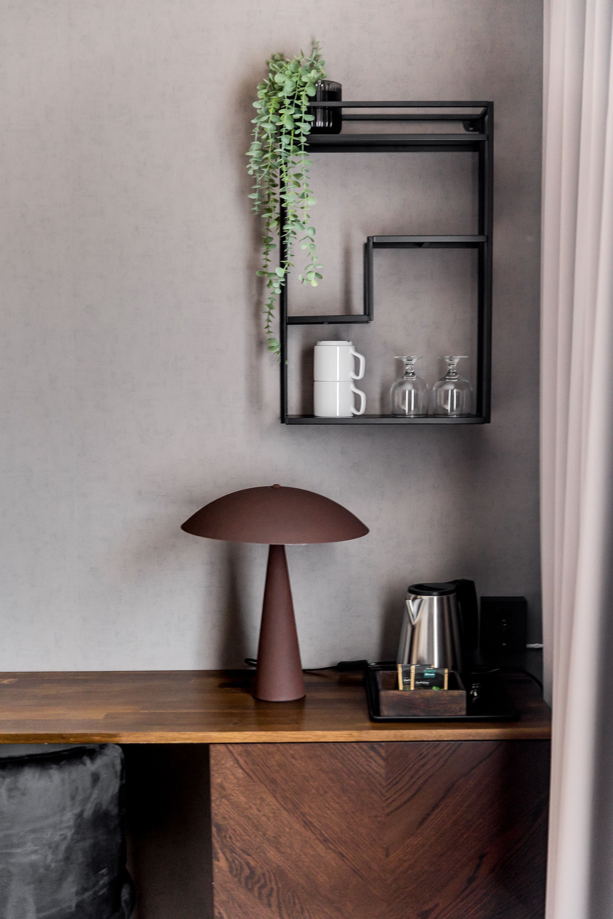Una habitación moderna con un escritorio de madera, una lámpara marrón, una estantería negra con una planta, una taza blanca y una cafetera.