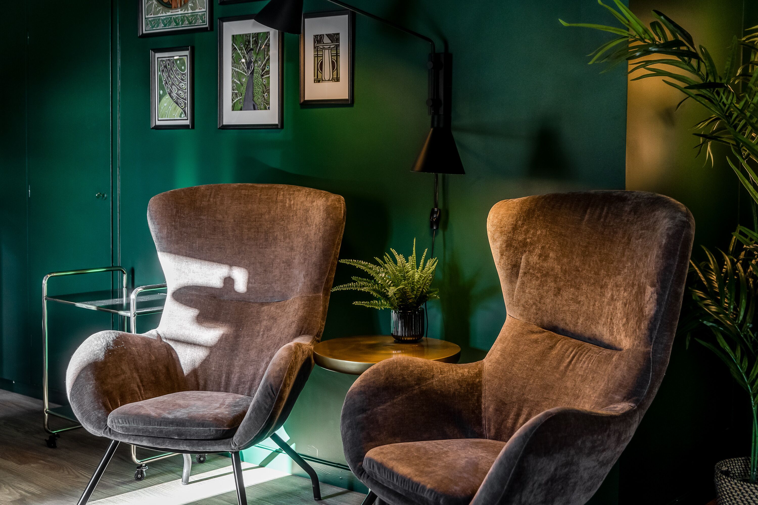 Two brown armchairs, a small round table with a plant, and a wall-mounted lamp in a room with green walls and framed artwork.